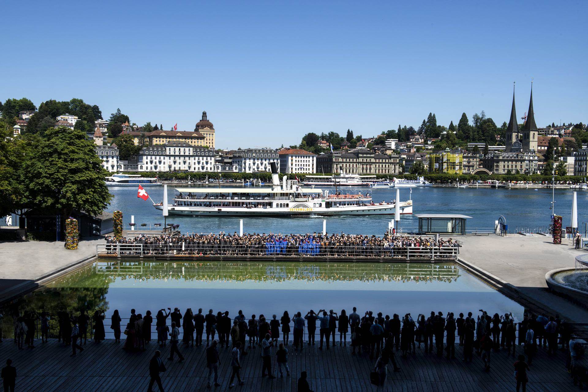 BHMS graduation on the Lucerne lakefront — graduates lined up above the reflecting pool as a paddle steamer passes behind them