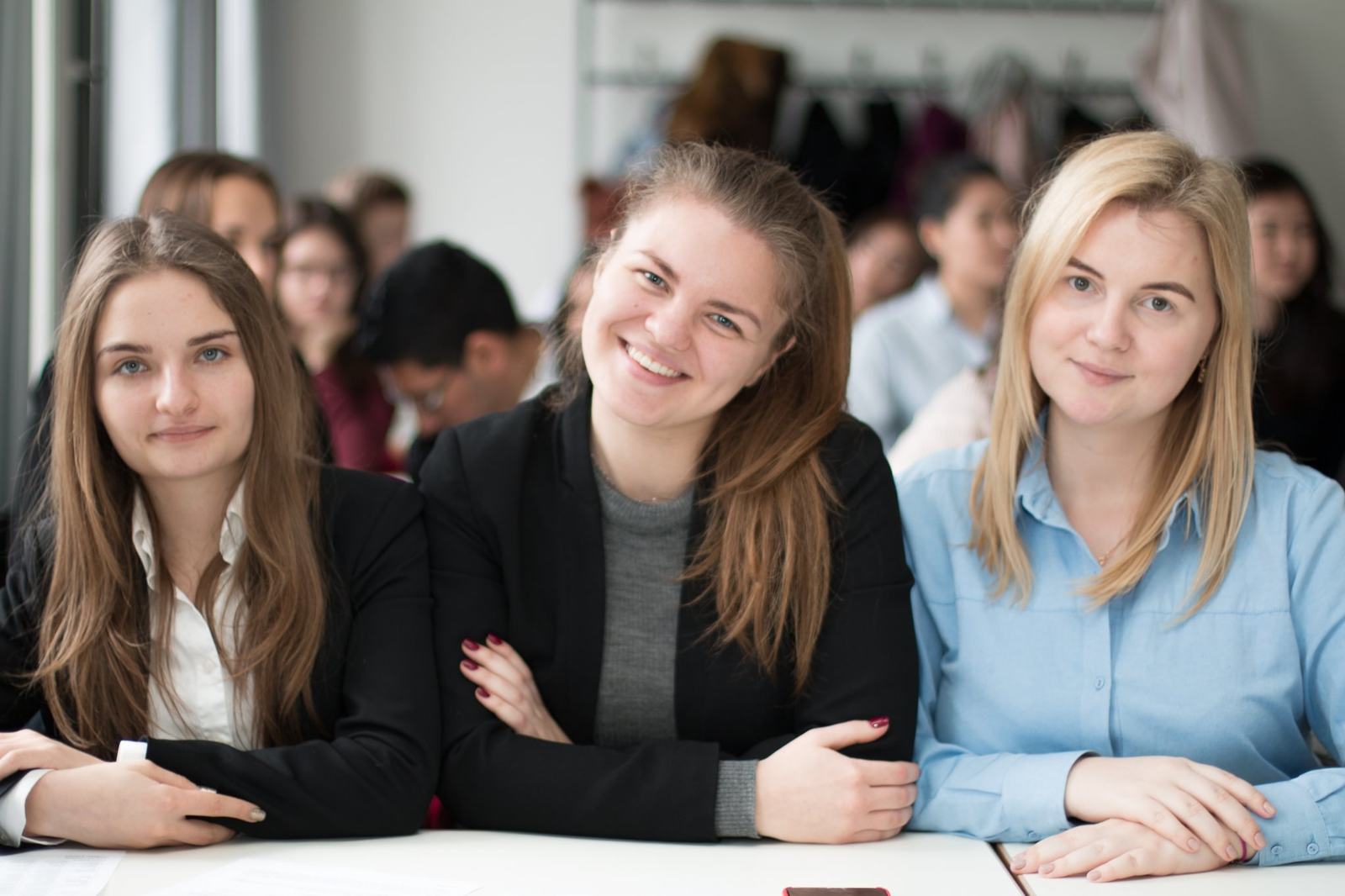 Three BHMS students in business dress at a classroom desk, classmates working behind them